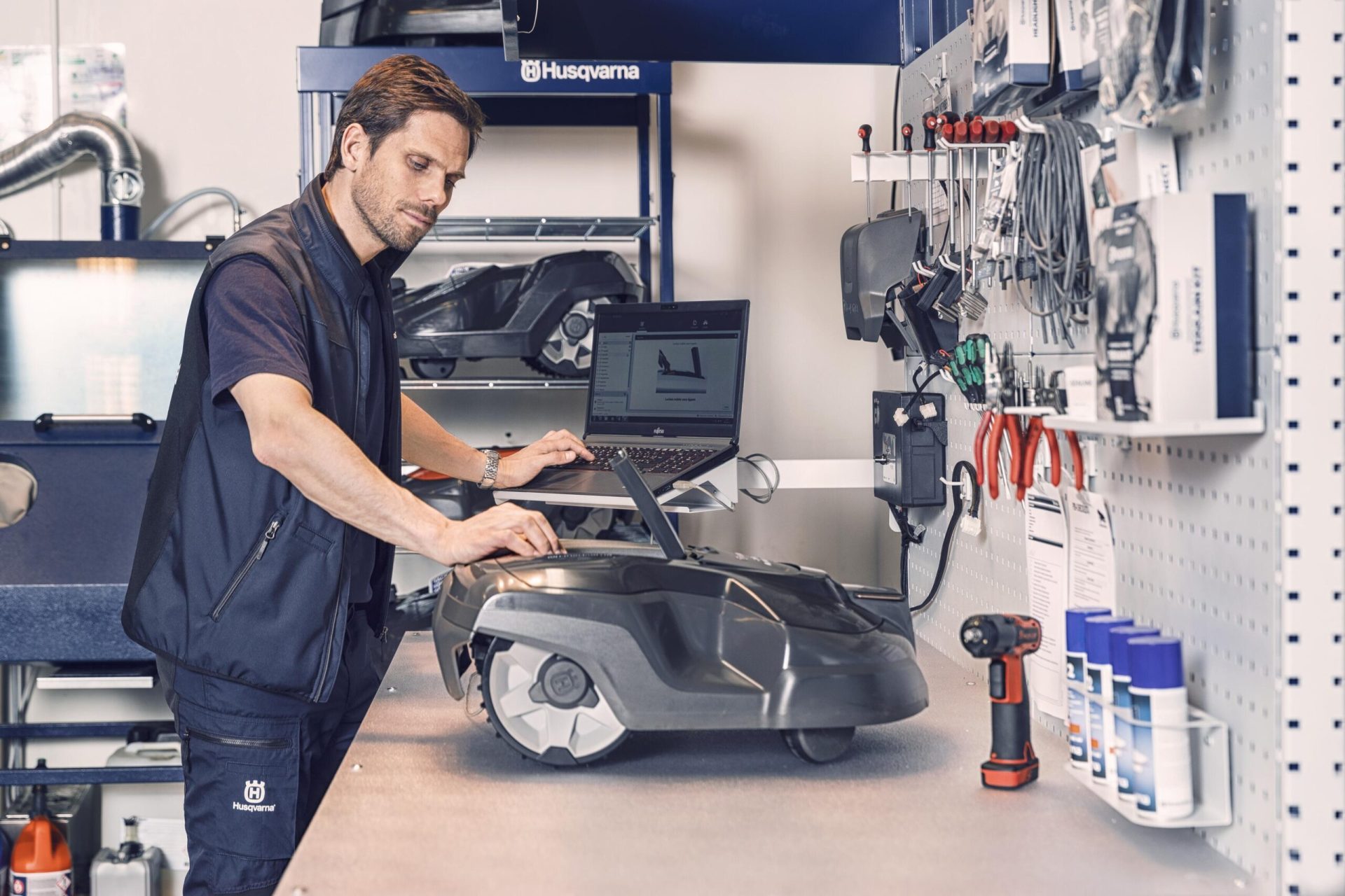 A man works on a auto mower in a workshop with a laptop and organized tools nearby.
